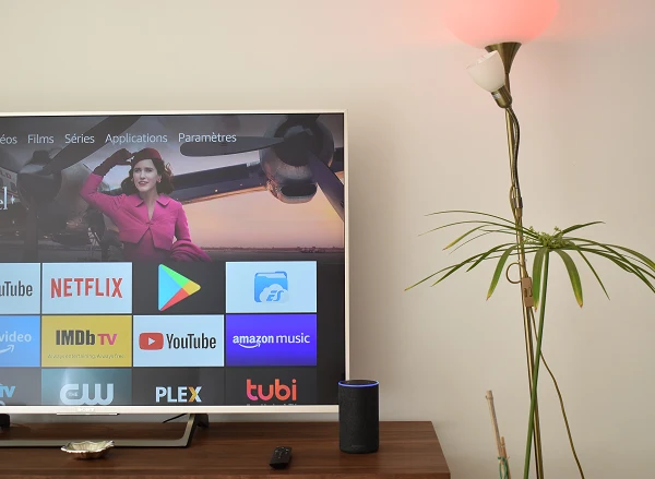 Amazon Alexa allume une ampoule. Derrière une télévision montrant l'interface d'Amazon Fire TV
